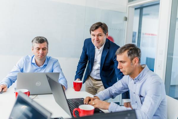 Three male consultants in a meeting room. Two of them working at their laptops, the thirs one looking friendliy into the camera